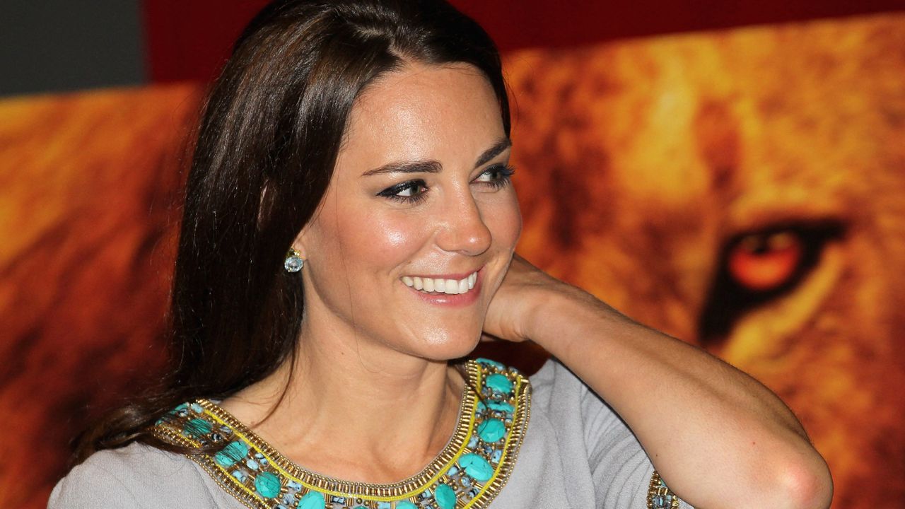 Kate Middleton wearing a gray dress with turquoise details on the neck smiling and resting her arm on her shoulder with a photo of a lion behind her in 2012