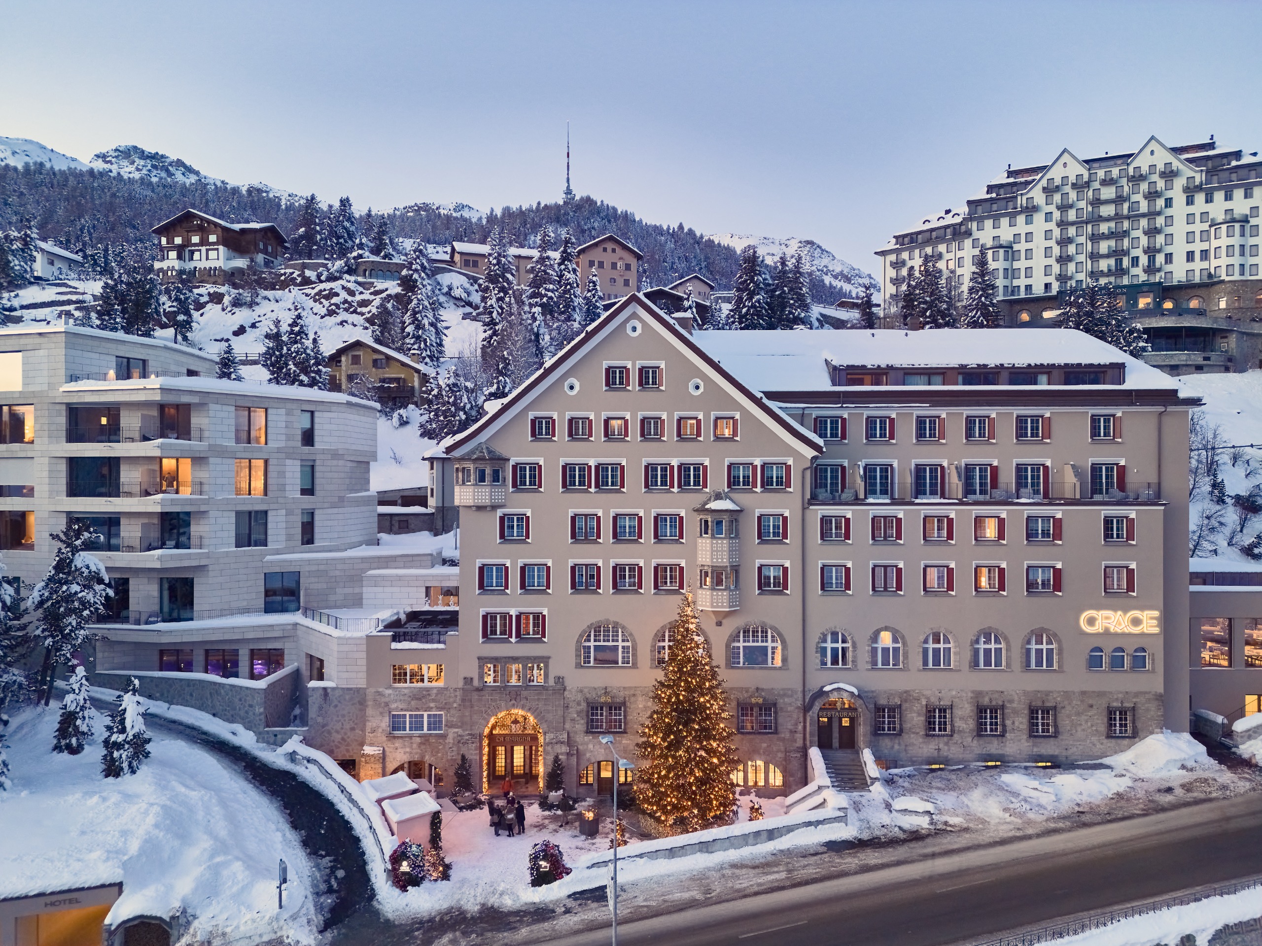 The exterior of a hotel in wintertime. The beige building with white windows and red shutters is surrounded by snow
