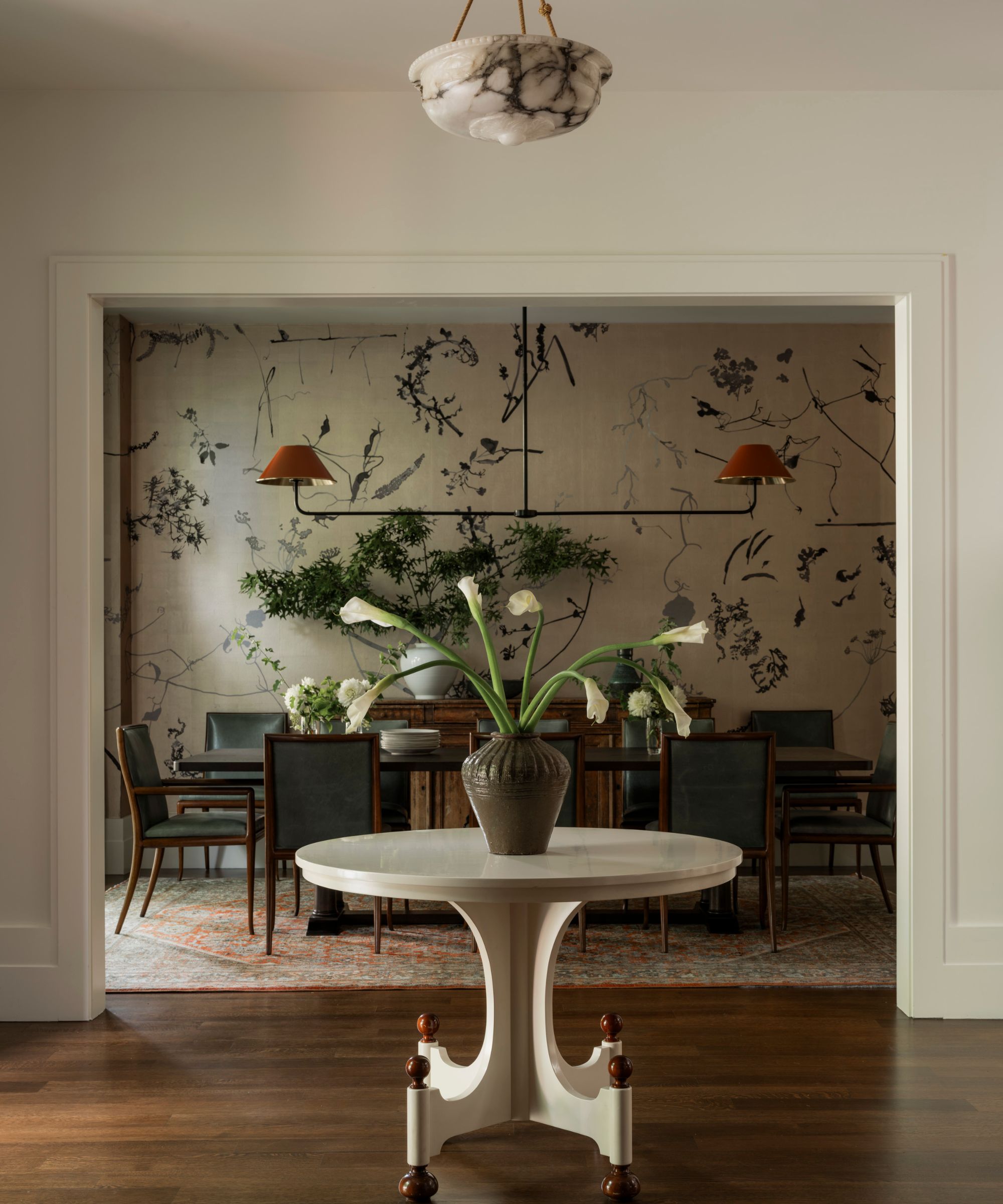 A view through a white doorway into an elegant dining room. In the foreground, a white pedestal table holds a vase with calla lilies. The dining room features a dark wood table, leather chairs, and a statement wall with a hand-painted botanical mural in black ink on a neutral background.