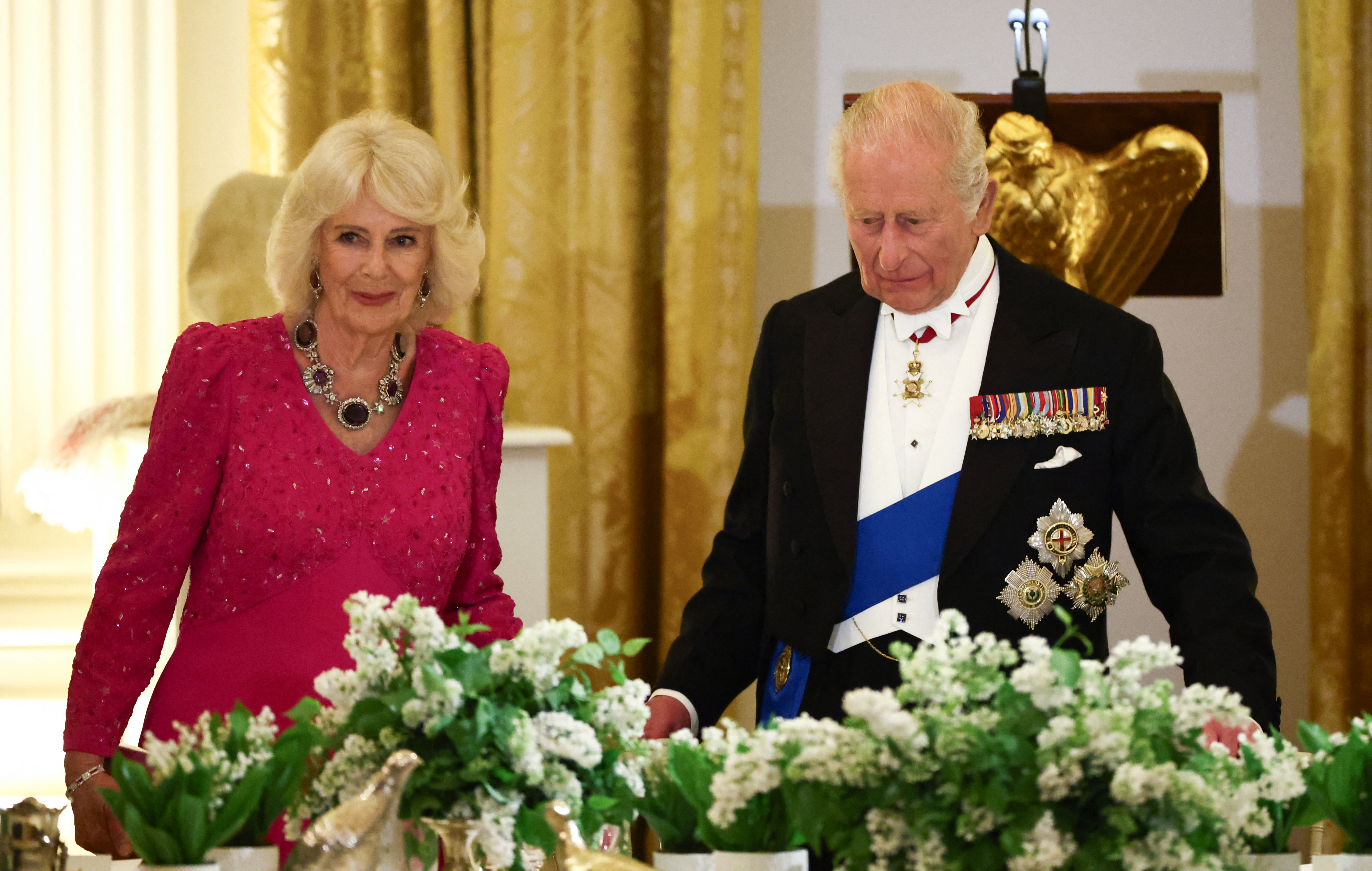 Queen Camilla wearing a pink gown standing next to King Charles at a banquet