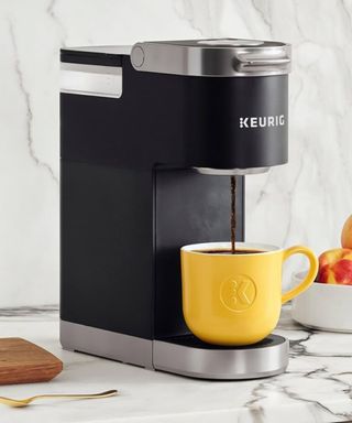 A yellow coffee beneath a Keurig K-Mini on a marble countertop against a white wall.