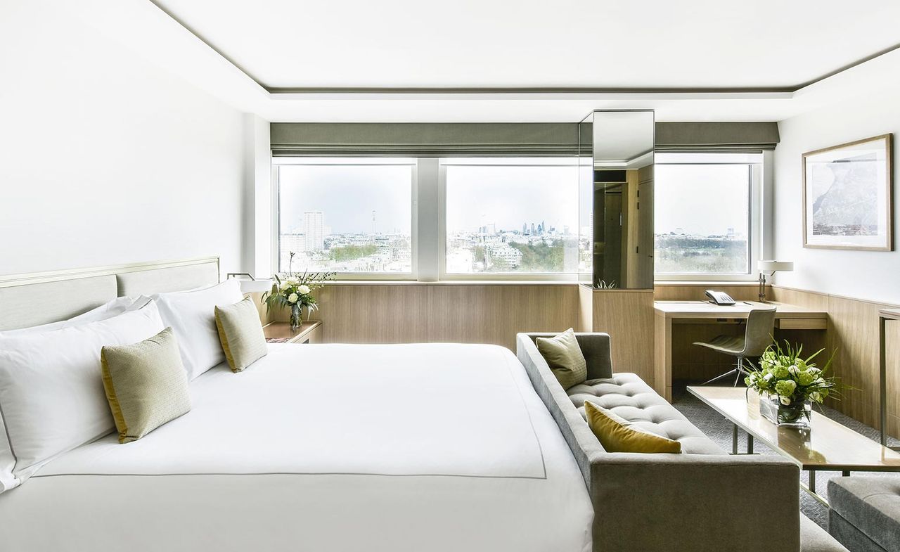 A room in the Royal Lancaster London. Three windows that go from wall-to-wall let in a lot of light. A large bed with a light gray headboard and white linen sits against the wall, with a gray couch and a coffee table in front of it.