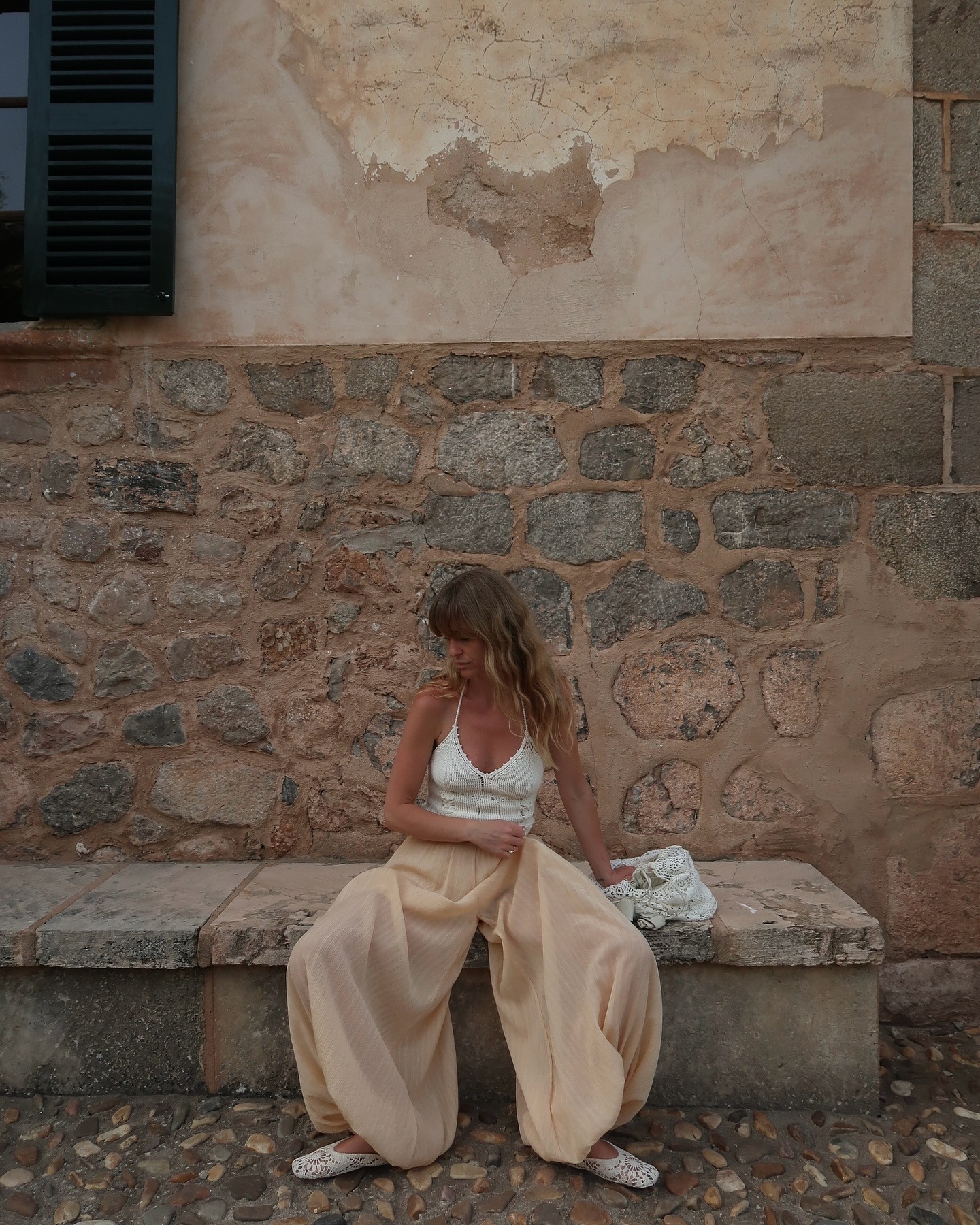 Fashion person Jeannette wears a crochet halterneck top, beige genie pants and crochet mules. She is sitting on a stone bench, in front of a stone wall, in a recent image from her Instagram.