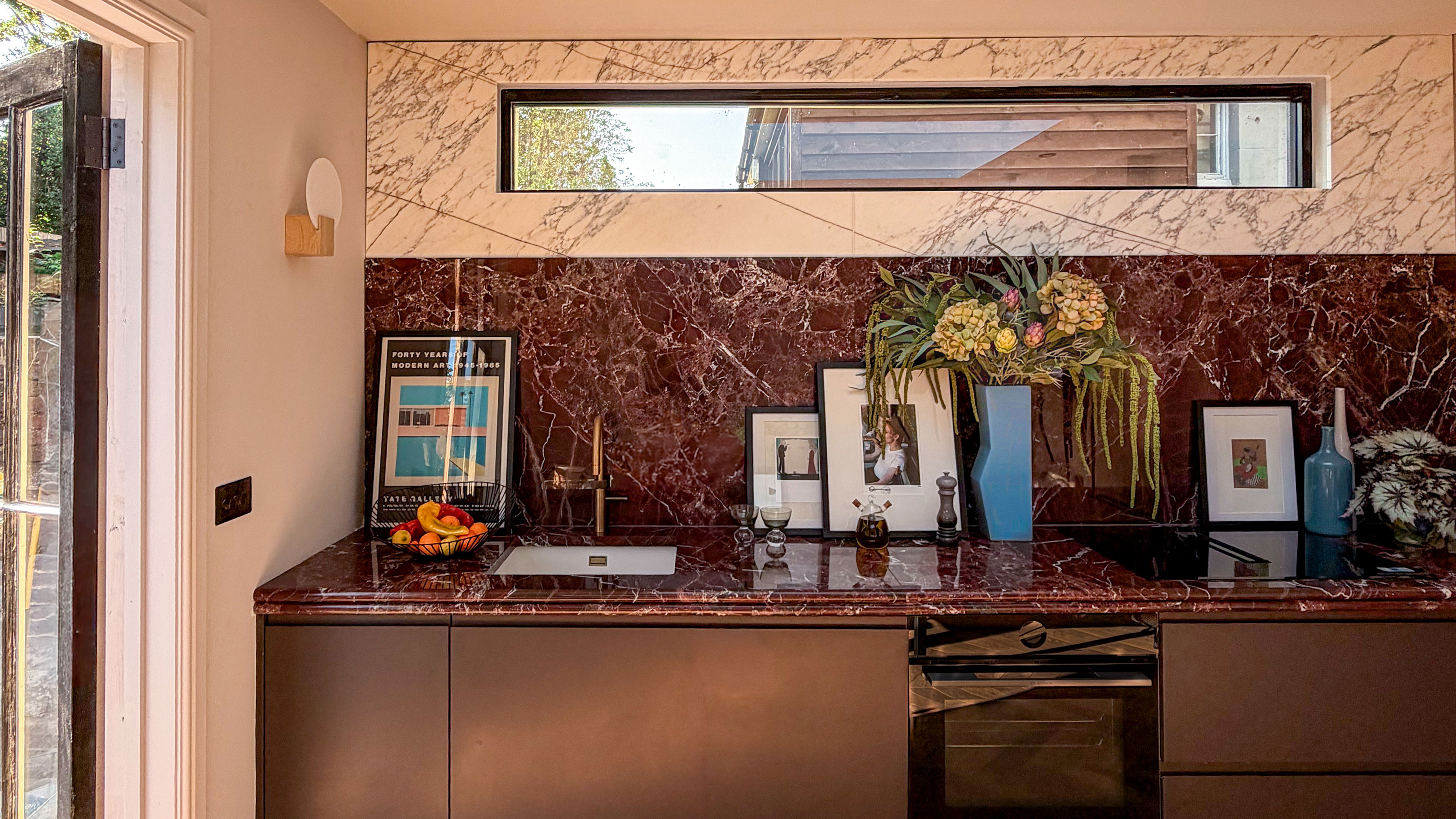 Kitchen with red marble worktops and splashbacks, topped with white marble. Dark red cabinetry. High narrow window. Photographs and vases on countertop