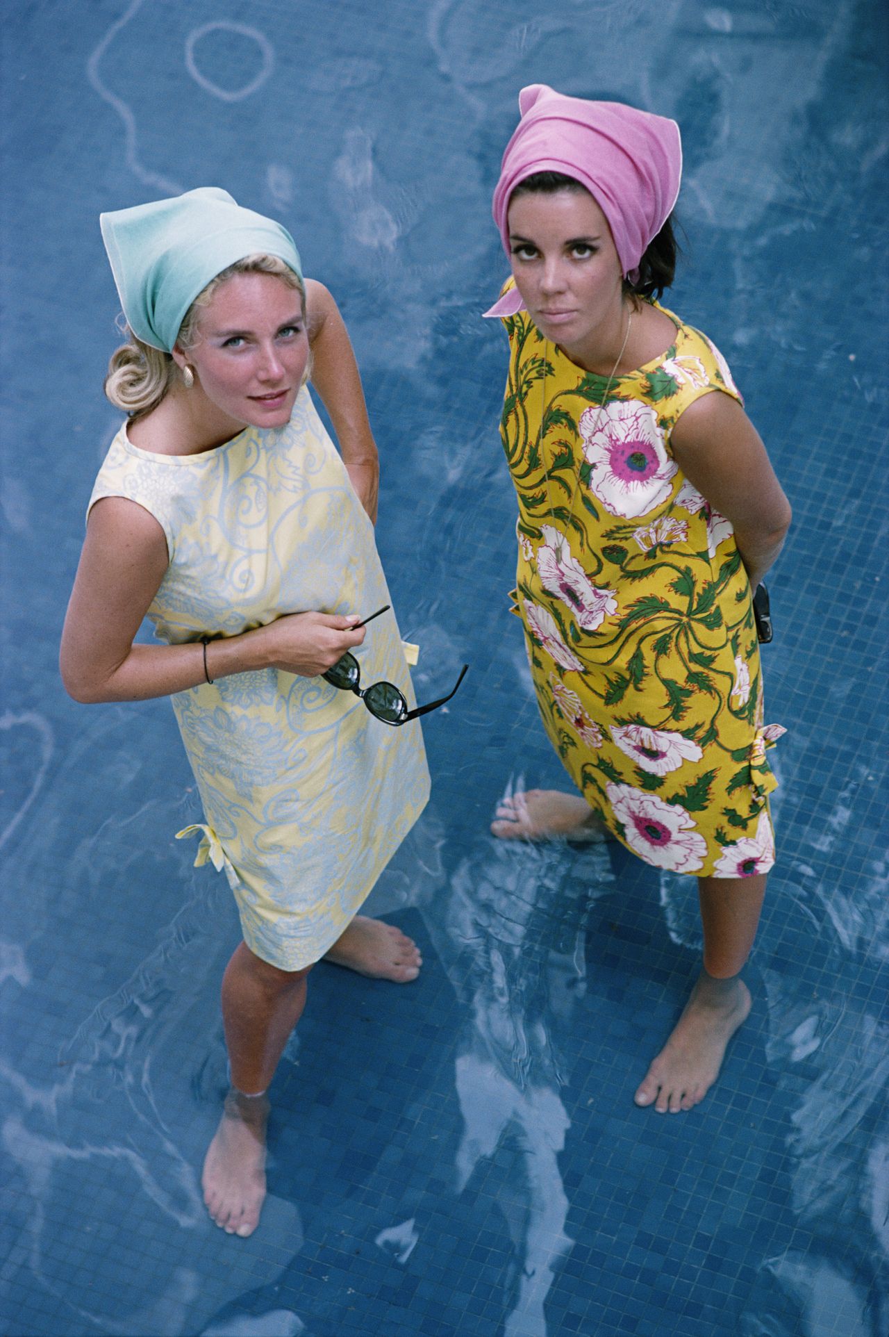 Two women in brightly-coloured, floral sun dresses, standing in ankle deep water, photographed from above