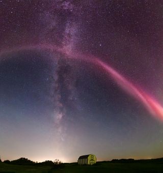 a long red stream of light arches over the night sky, in the background is the milky way appearing as a thick band of stars running from the top of the image down to the horizon.