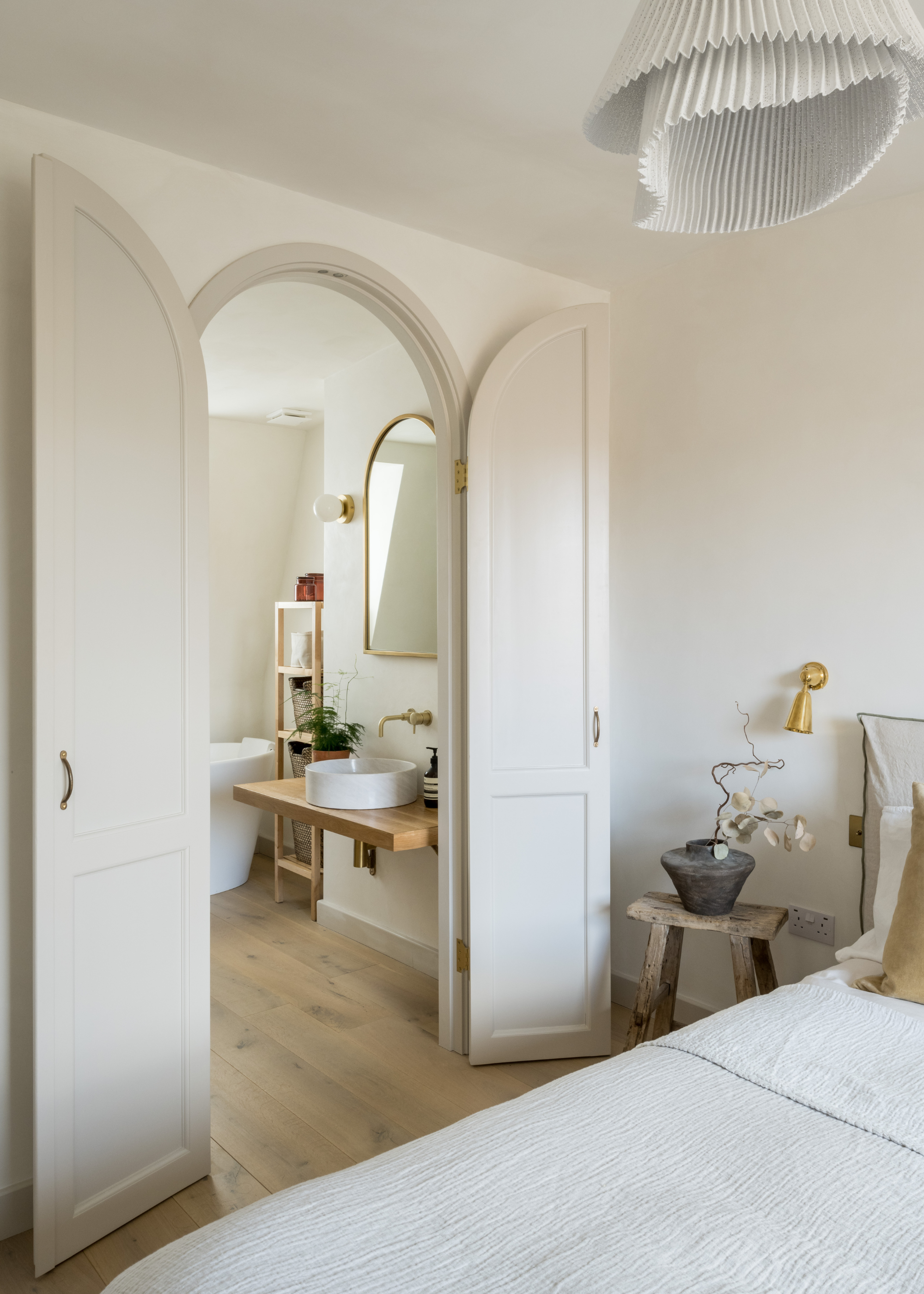 A minimalist bedroom with a low bed, a natural bedscape, a side wood stool with a vase of stems, gold wall sconces, and a pleated pendant light leading to a pared back bathroom