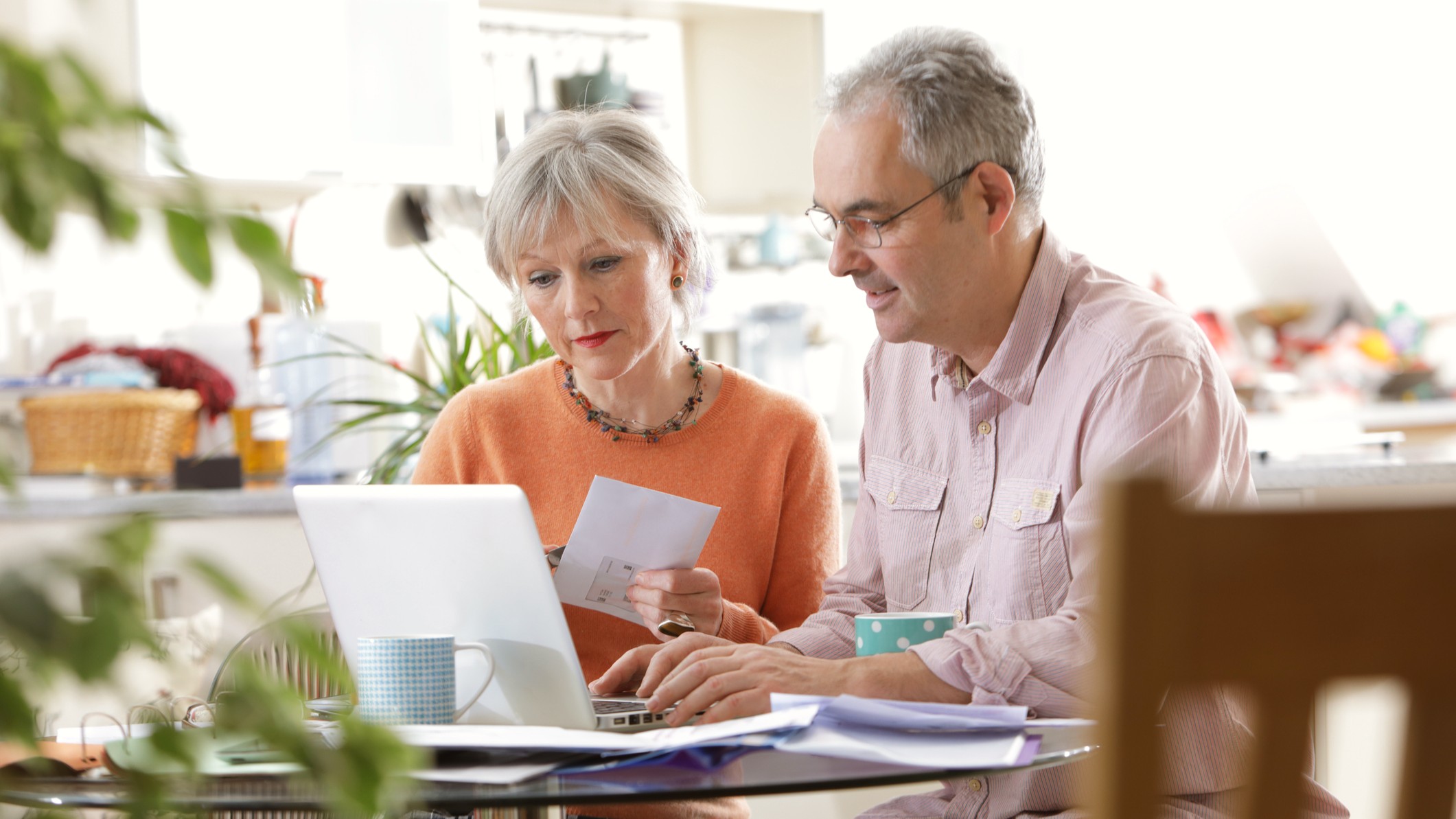 An older attractive couple sits at the kitchen table, looking at a laptop. They appear to be discussing financial matters. 
