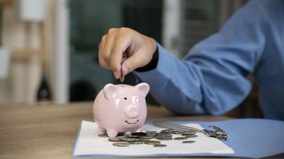 A man in a blue dress shirt drops a coin into a piggy bank, only his hand and arm showing.