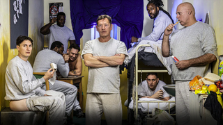 British actor and Banged Up participant Sid Owen stands, arms crossed, in a busy prison cell surround by lags. 