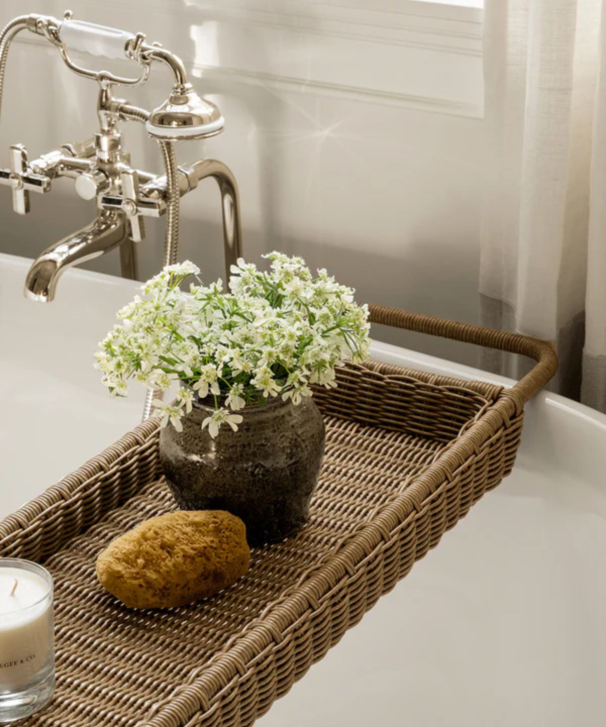 A close-up of McGee &amp;amp; Co.'s rattan bath caddy, displaying a small vase of flowers, a luffa, and a candle