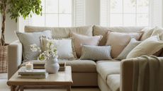 A white corner sofa with piles of pastel cushions on it, a wooden coffee table in front