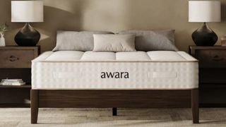 The image shows a pale beige mattress on a wooden bed frame, placed within a neutrally decorated bedroom. 