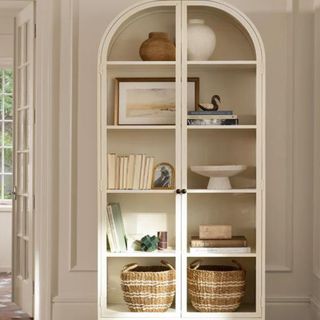 An arched white cabinet with glass doors and two wicker baskets on the bottom shelf