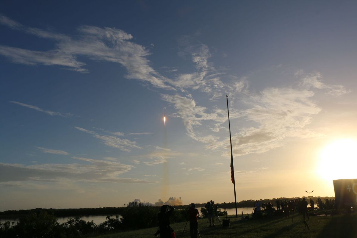 A rocket launches in the distance against a blue sky with some clouds.