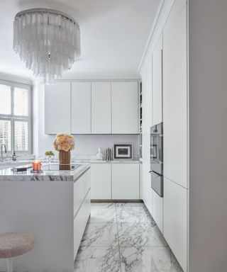 All white kitchen with white and gray marble countertops and flooring, kitchen island with upholstered bar stool, large glass chandelier over island,