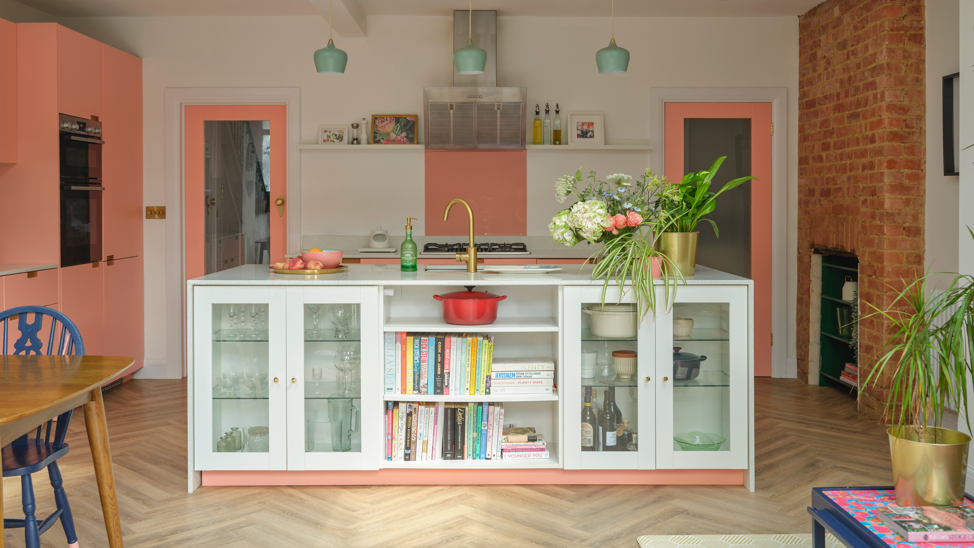 a colourful pink and white contemporary family kitchen with a trio of minute green pendant lights, herringbone wooden flooring, and exposed brick chimney breast and a white glazed door kitchen island