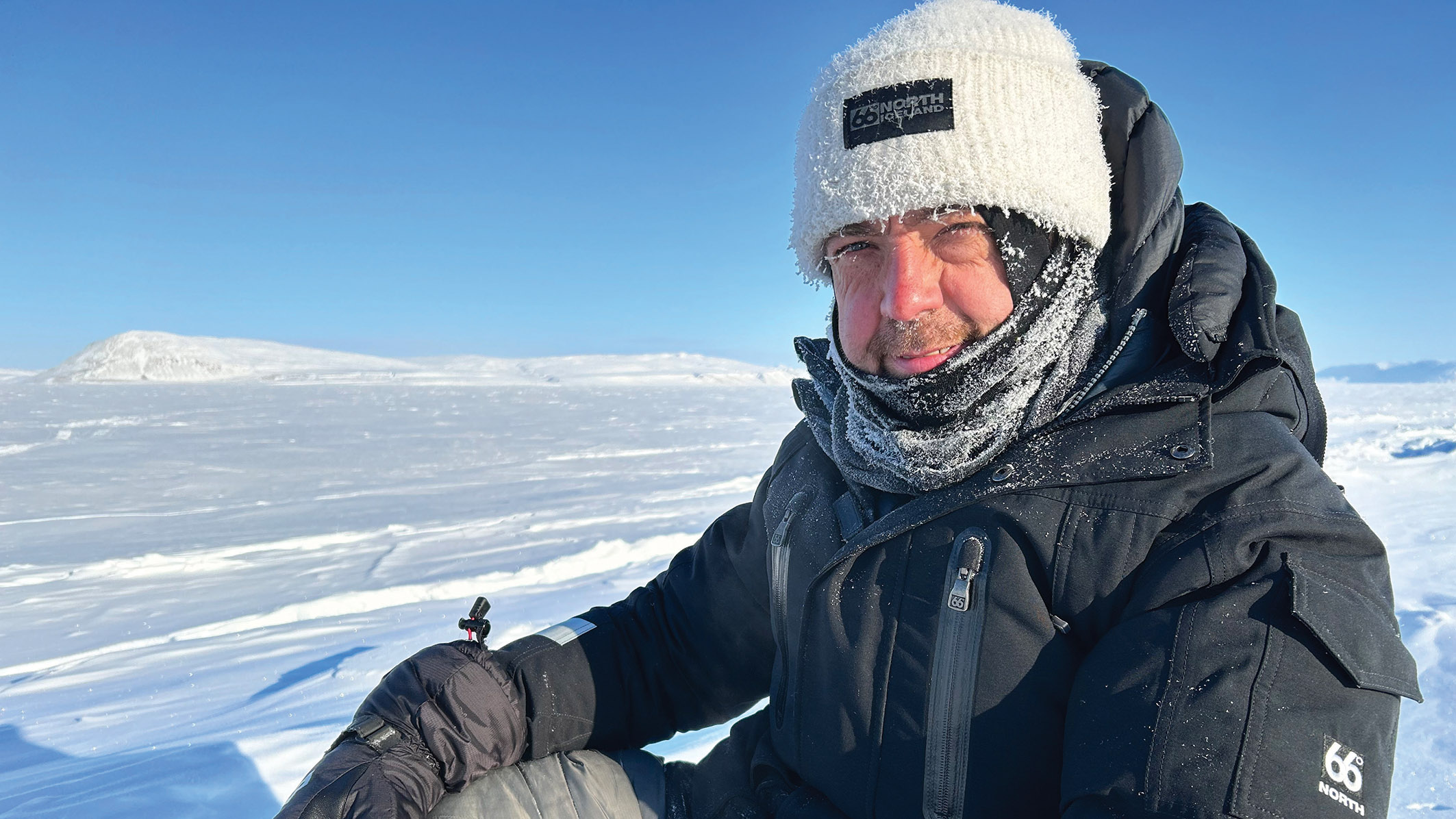 A person in a warm black jacket and white beanie sits on snow, surrounded by vast, icy terrain under a clear blue sky