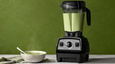 A Vitamix propel series 750 blender with green soup inside, against a green background. There is a small white bowl of green soup beside it. 