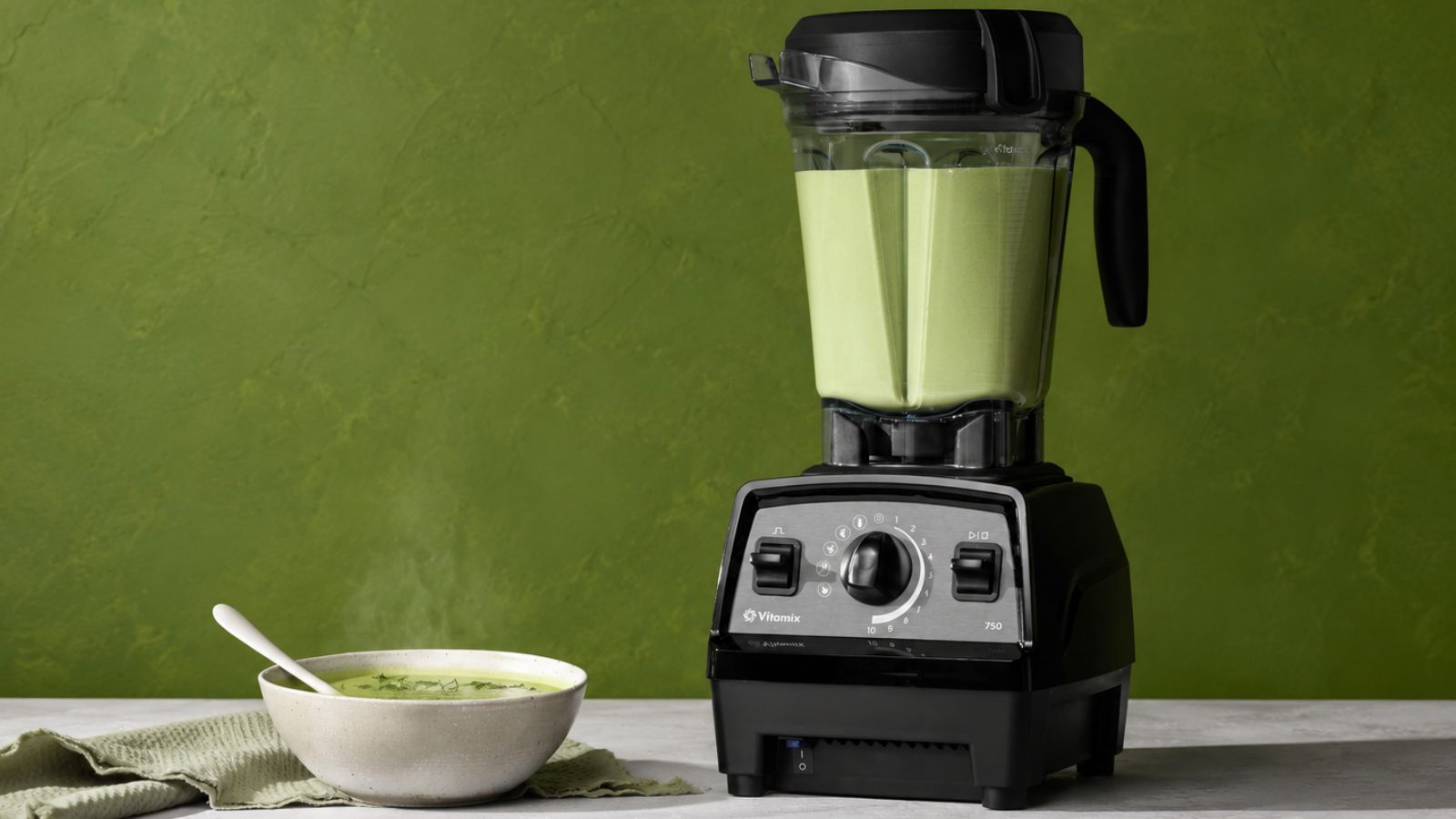 A Vitamix propel series 750 blender with green soup inside, against a green background. There is a small white bowl of green soup beside it. 