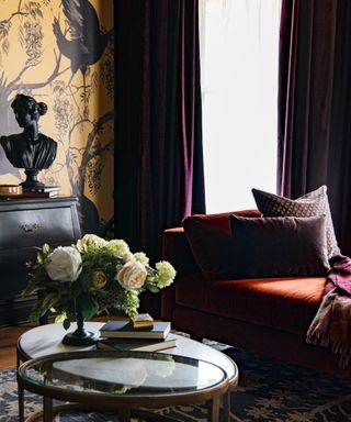 living room corner with dark red sofa, glass coffee table with green and white flowers, yellow patterned wallpaper and dark purple curtains