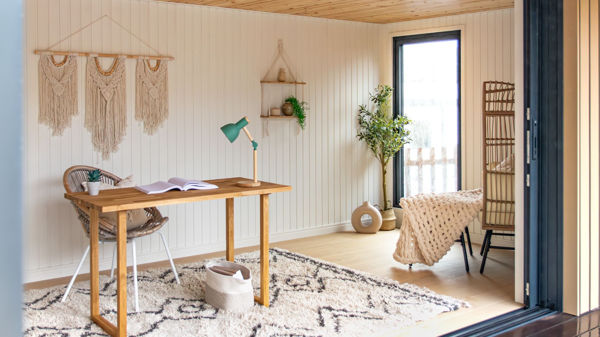 A light-filled garden office interior with white tongue-and-groove walls, a pine ceiling, oak desk, rattan chair, Berber rug, macramé wall hangings and bifold doors.