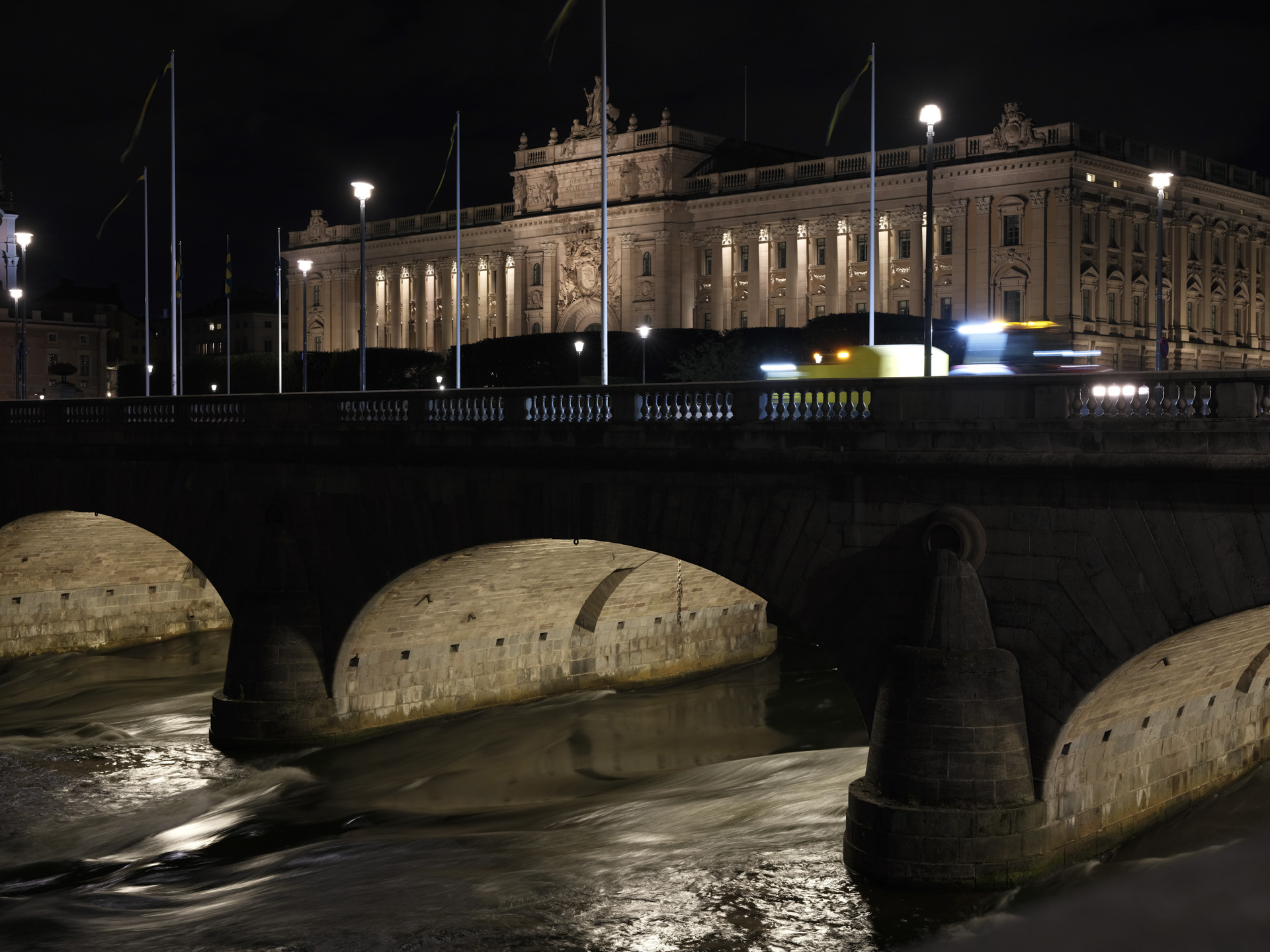 Night cityscape of a grand building framed by a bridge and river with a 0.5 seconds slow shutter speed blurring the water, shot handheld