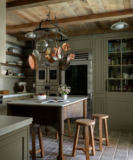 A baking kitchen with green wall tiles, green cabinets, and green wall shelves. A freestanding wooden prep table sits in the middle of the room with a ceiling-mounted pot rack suspended above