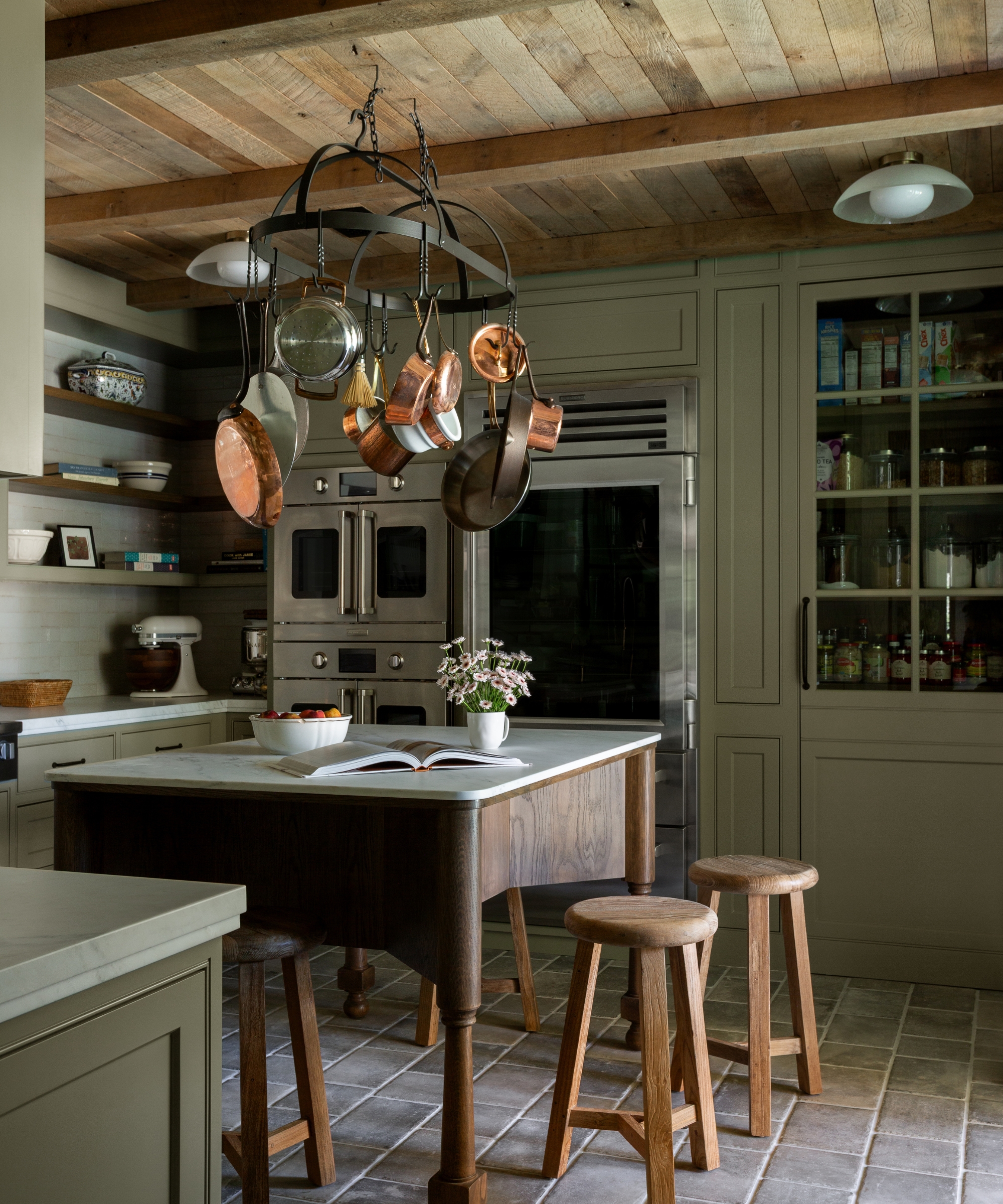 A baking kitchen with green wall tiles, green cabinets, and green wall shelves. A freestanding wooden prep table sits in the middle of the room with a ceiling-mounted pot rack suspended above