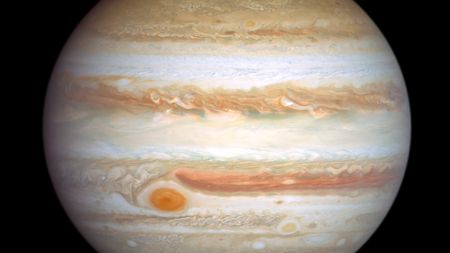 A tan, gaseious Jupiter hangs in black space, layered like a cross section of geological survey, a swirling red storm at its lower left.