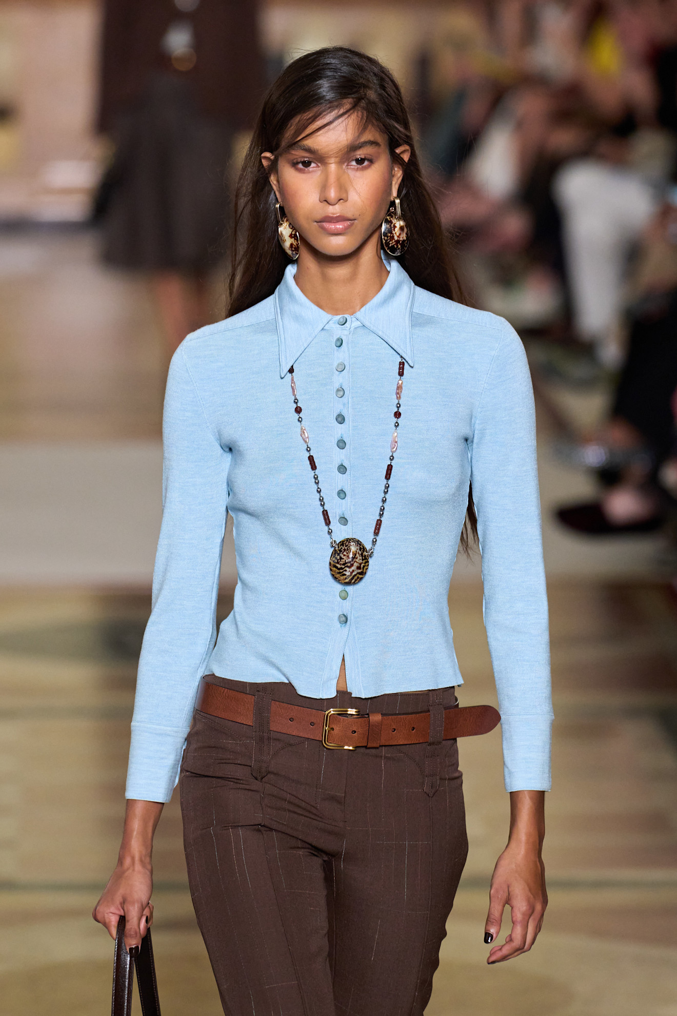a woman wearing a blue knit top with brown pants and a pendant necklace