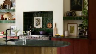 Kitchen with dark red wood cabinets, yellow tiled island, dark green stone countertops and zellige splashback tiles, and shelving lined with objects