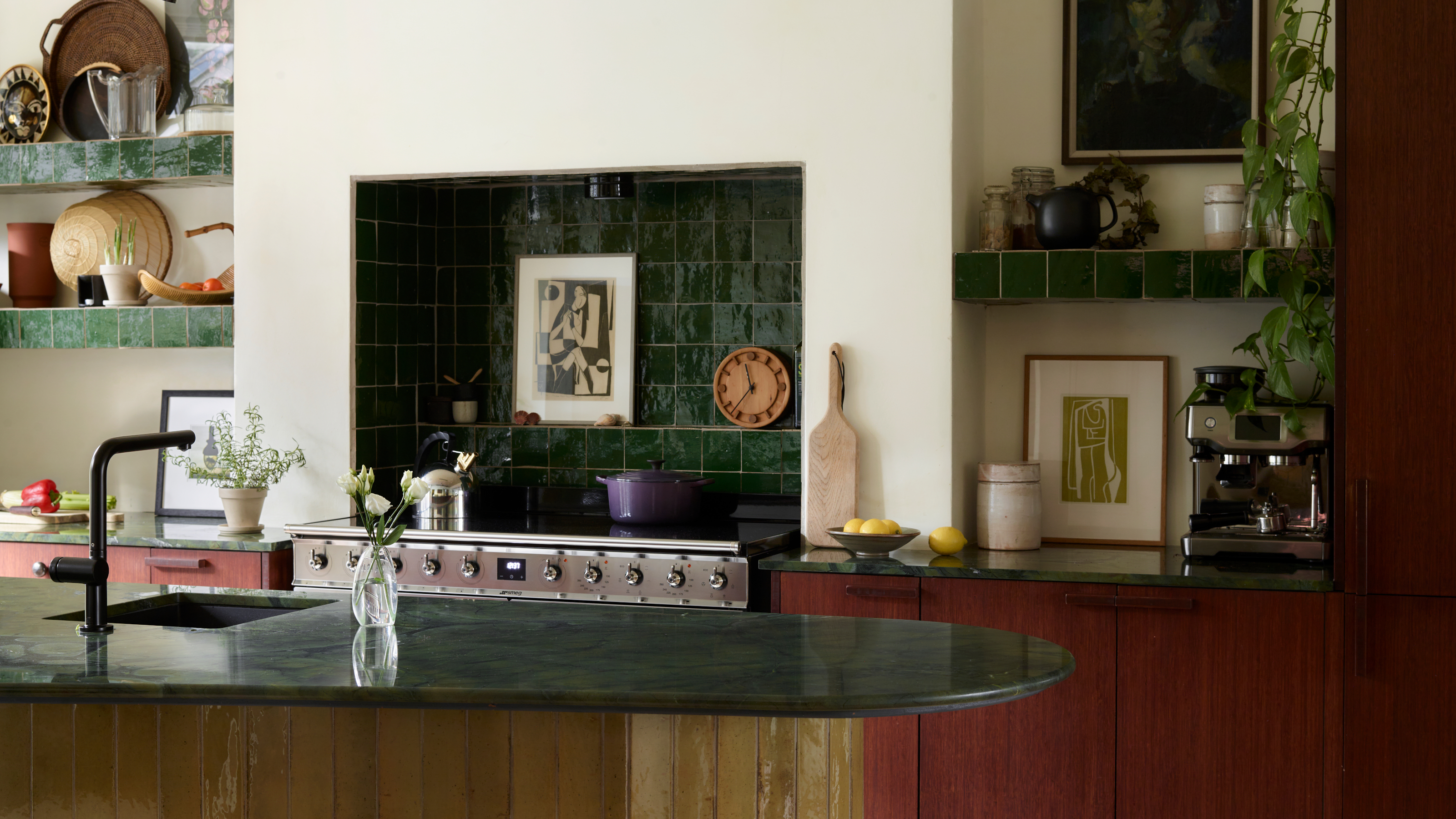 Kitchen with dark red wood cabinets, yellow tiled island, dark green stone countertops and zellige splashback tiles, and shelving lined with objects