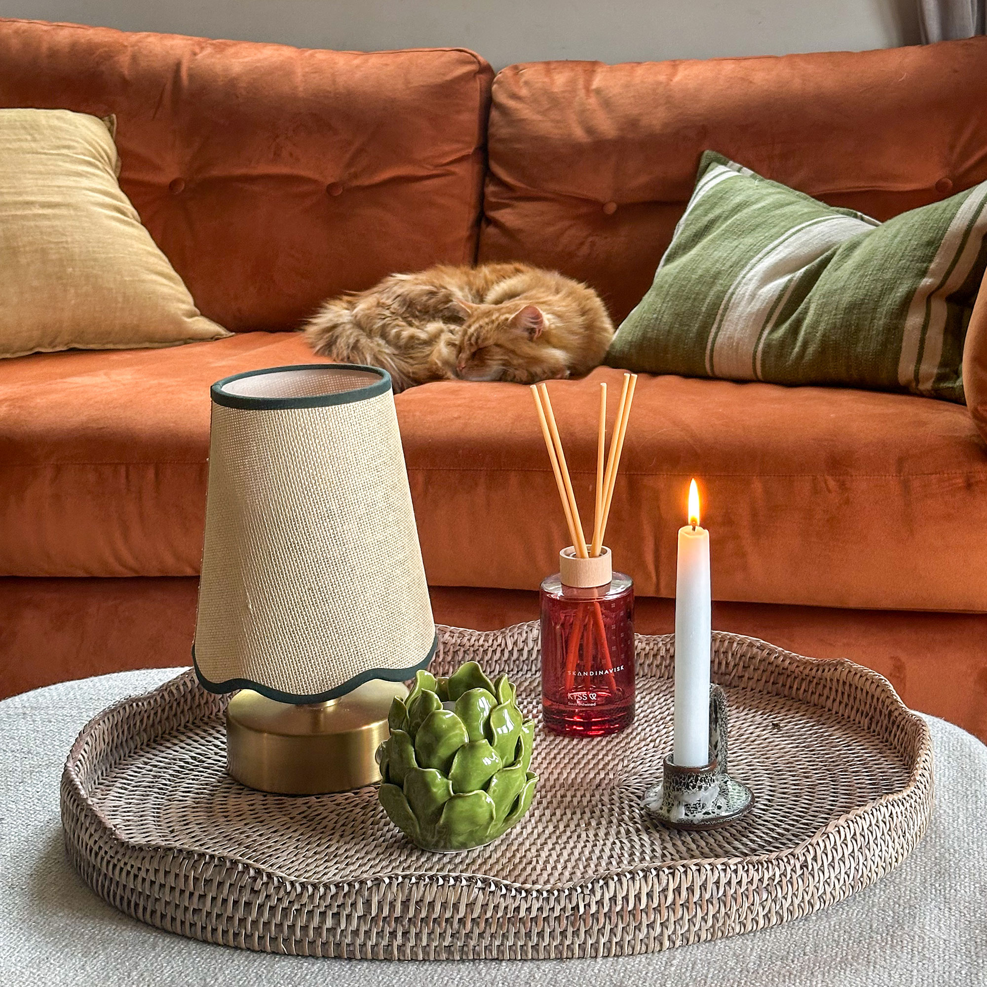 Rattan tray holding wireless lamp, candle and reed diffuser in front of orange sofa with cat
