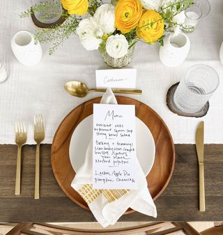Wooden plate setup with gold-plated cutlery and table card with surrounding white and yellow flowers