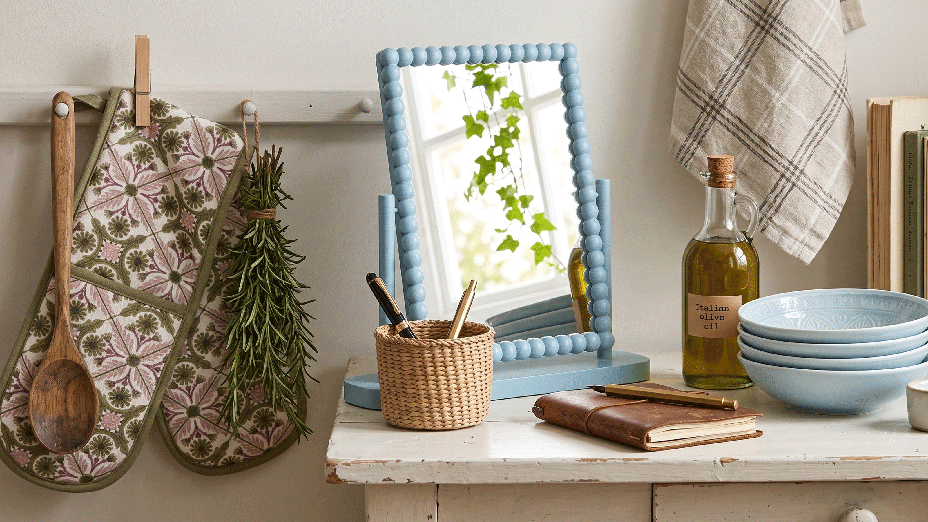 a kitchen counter or table with a blue-framed mirror, with a stack of blue bowls, a wooden spoon and a patterned oven mitt with a bunch of rosemary