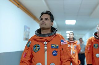 three astronauts in orange flight suits walk down a white hallway.