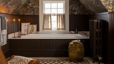 moody loft guest bathroom with dark wooden brown panelling and bath panel, small window with cafe curtain, botanical wallpaper, decorated with a rug, stool, artwork, candles and a small chair