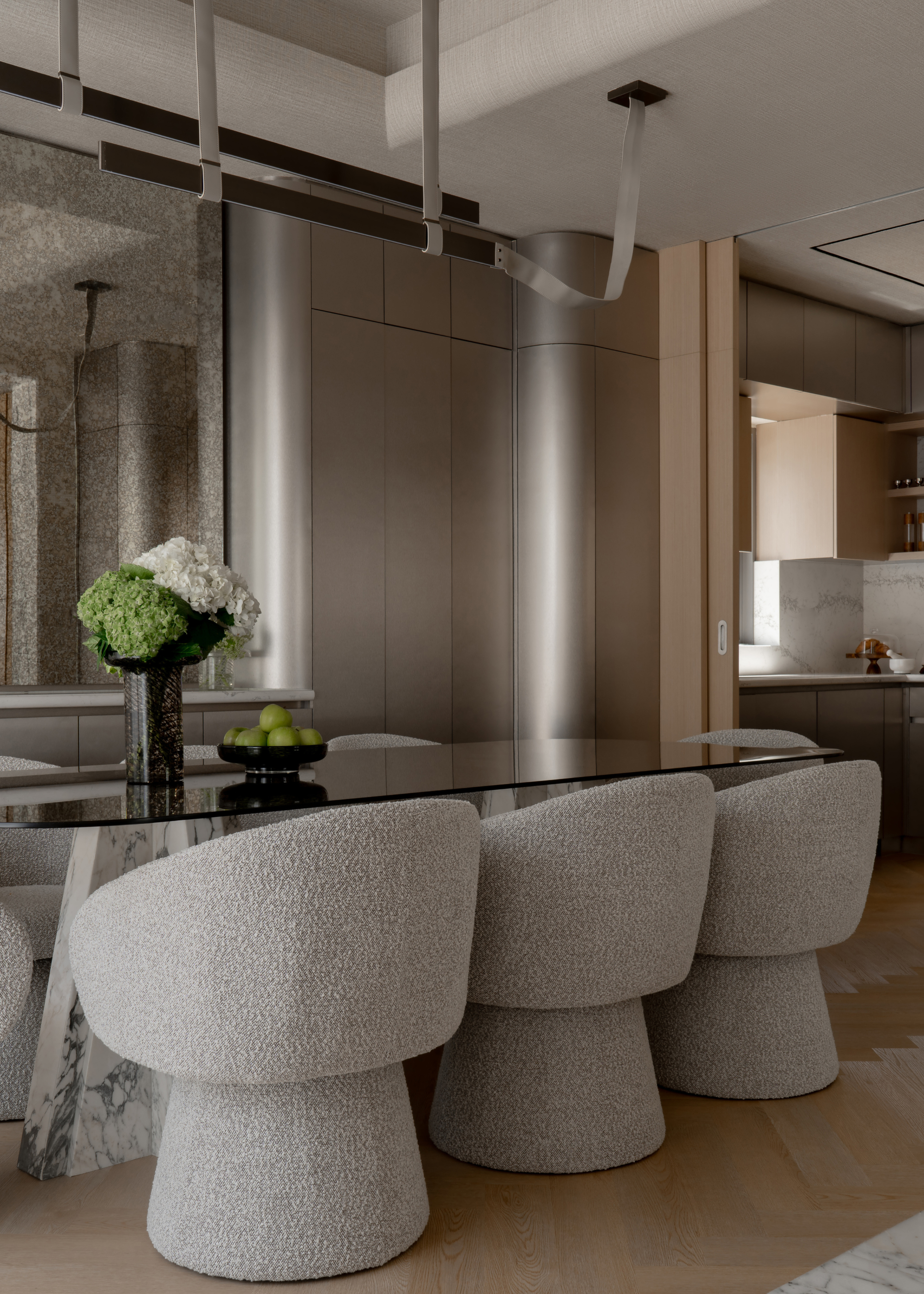 A dining room with a tinted glass table with stone legs alongside a set of gray chairs by an antique mirror wall and matte silver cabinets