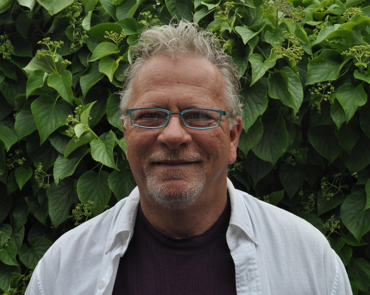 Headshot of man in white shirt and glasses in front of a hedge