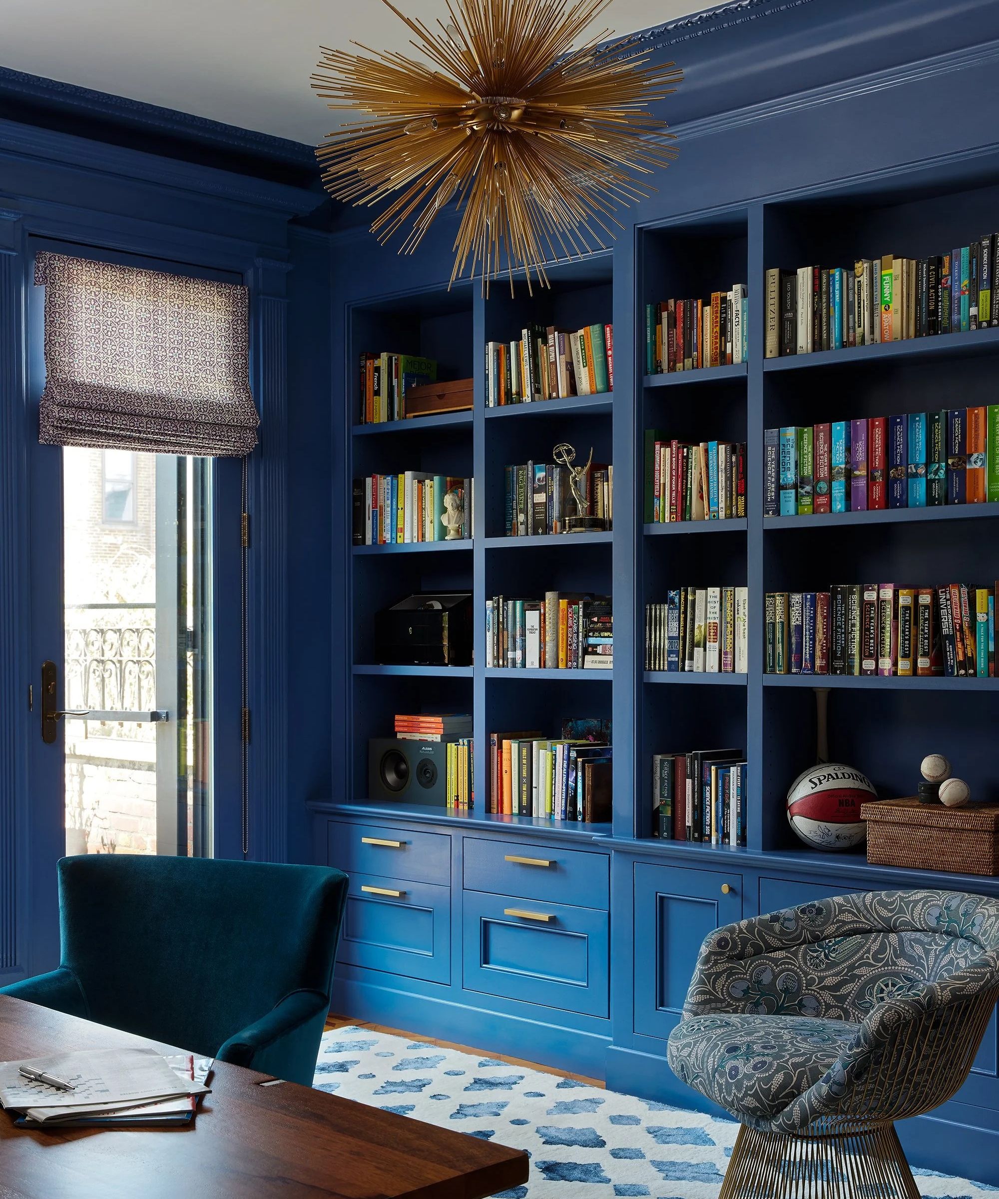 a bold home office with blue painted walls and shelves, shelves styled with books and other decor, and a bold gold chandelier from the ceiling