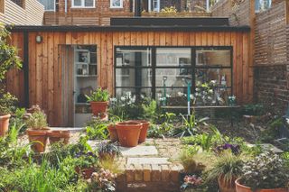 a wooden artists studio garden room , with shelves inside, and a full, lush paved garden outside with lots of potted plants and gardening essentials