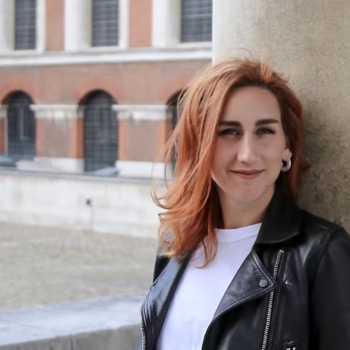 Image of Marta Balazs, a woman with red hair wearing a white t-shirt and black, leather jacket. She is standing outside in a building's courtyard.