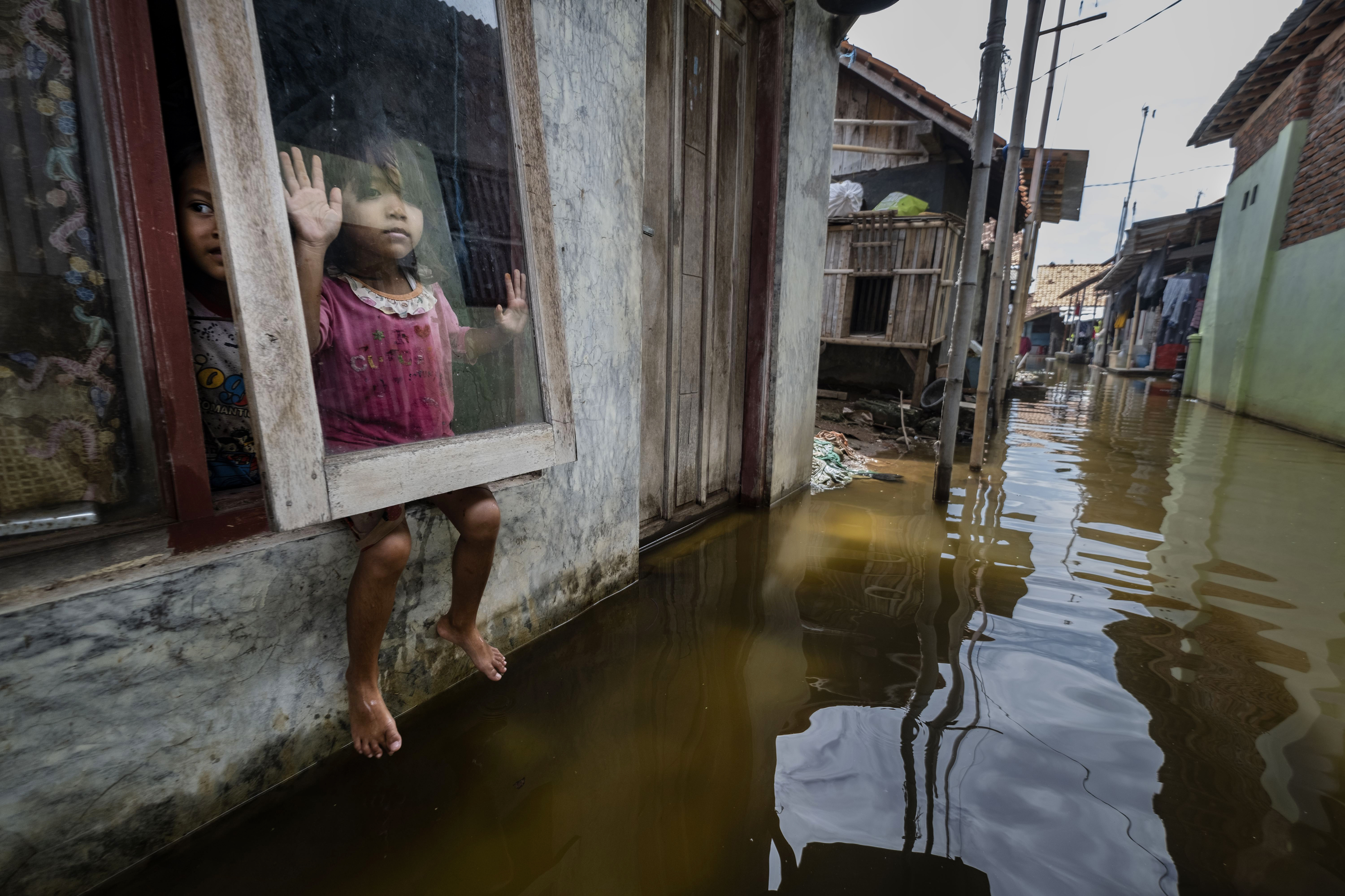 Two children look out from a window of their flooded home in Java, Indonesia, one pressing her hand against the glass and dangling her bare feet above the murky brown floodwater