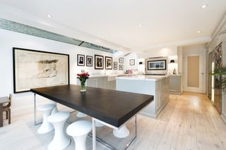 a modern and characterful L shaped kitchen extension - with a wide oak dining table and white stools, grey country-style cabinetry and large square island, and light wooden floor, with a large painting to the left