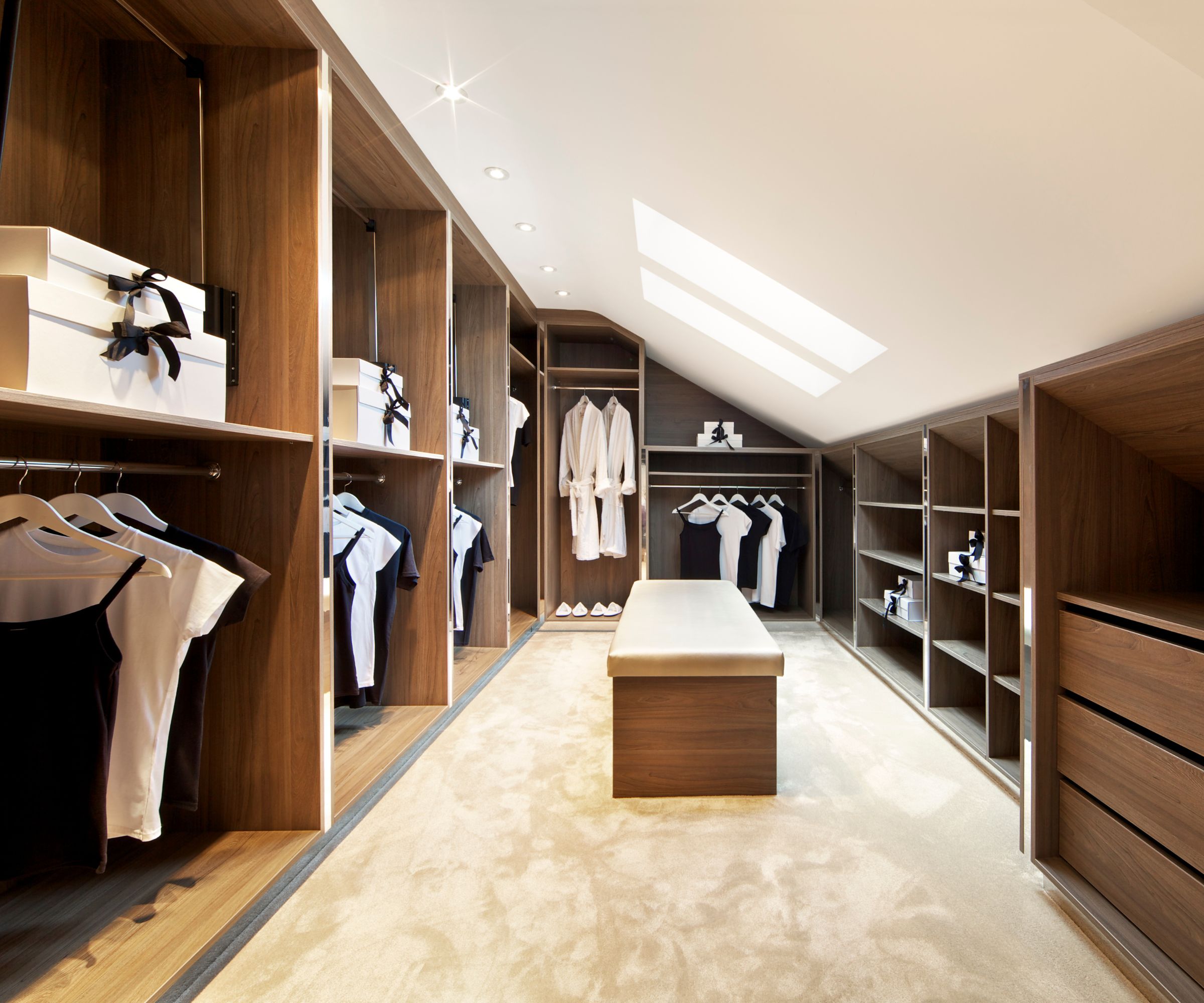A bright and spacious walk-in wardrobe in a luxury penthouse apartment with fully fitted walnut shelves, cupboards and drawers. A large suede covered ottoman stool sits in the centre. Clothes, boxes and bags have been used to dress this scene whilst the room is lit by a row of warm spot lights and two skylight windows.
