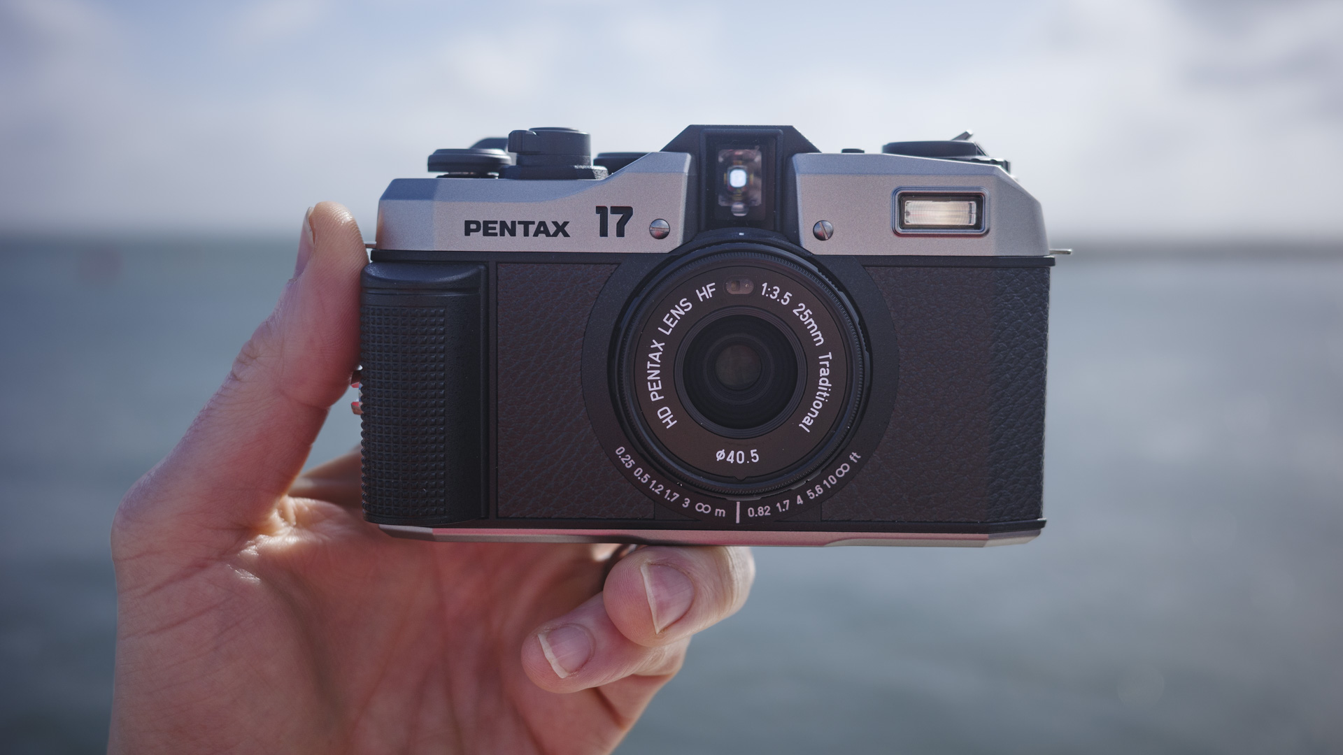 Pentax 17 compact film camera  front-on, in the hand  with ocean backdrop