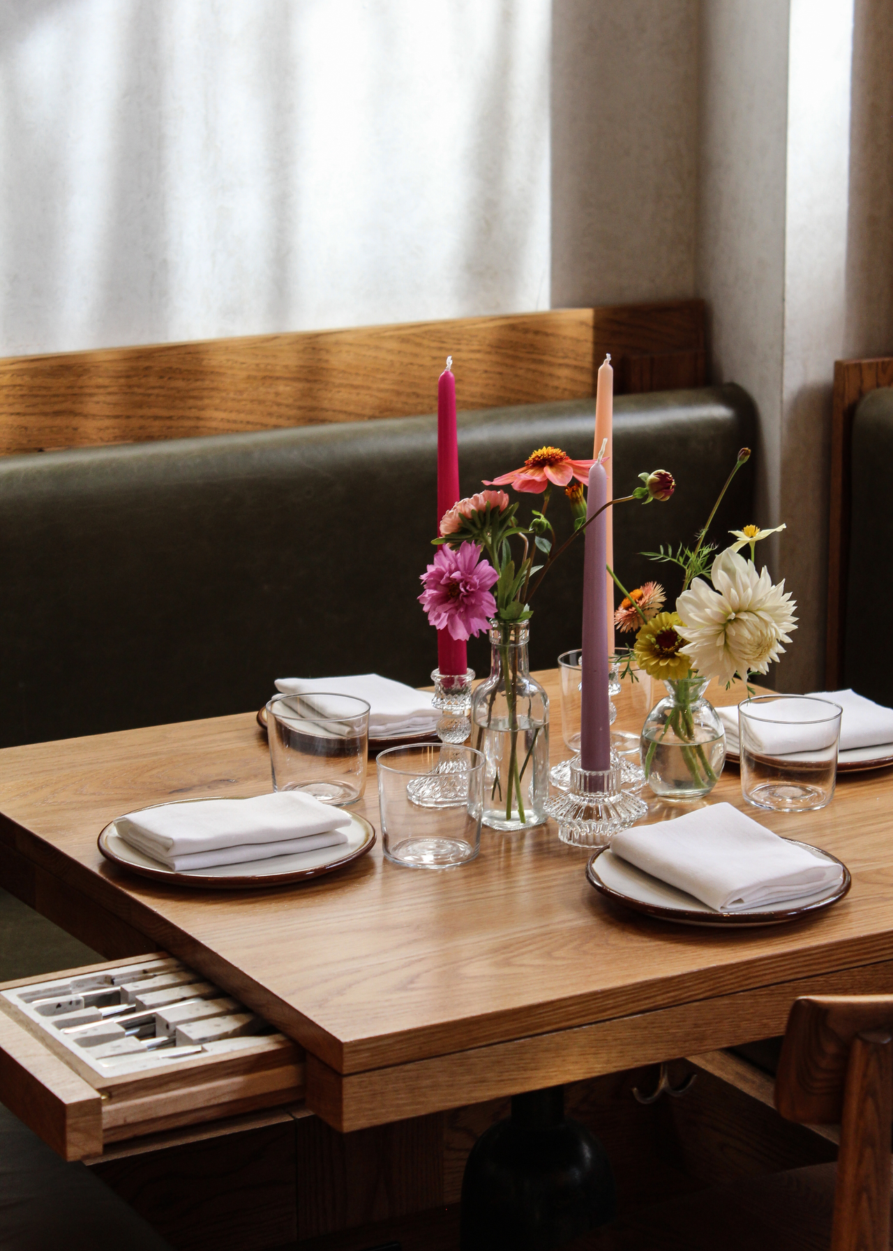 A restaurant with a wood dining table set with candles, plates, flowers and napkins alongside a storage drawer featuring a cutlery insert