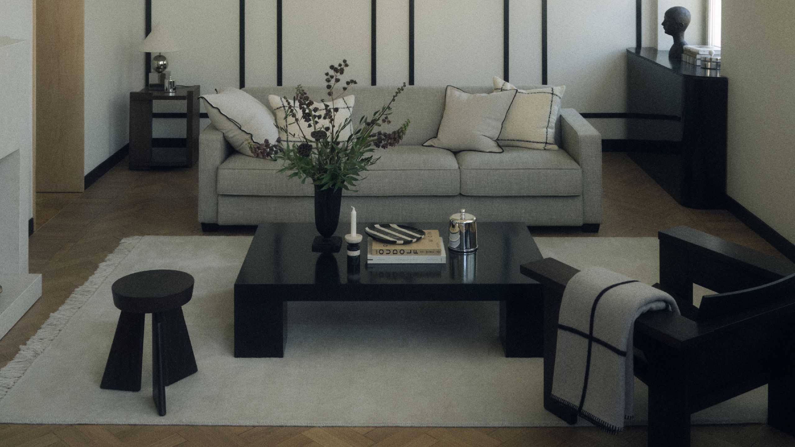 A monochrome living room with a gray sofa, cream rug and black coffee table, armchair and stool with a black console in the back right of the room by a window and black and white detailing throughout