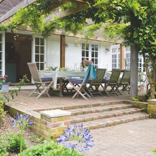 Wooden garden furniture on block paving patio area with steps under wooden pergola with climbing plants